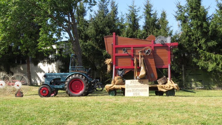 Un tracteur bleu se tient à côté d'une boîte en bois rouge artistiquement conçue. En arrière-plan, des arbres verts sont visibles, créant une atmosphère rurale.