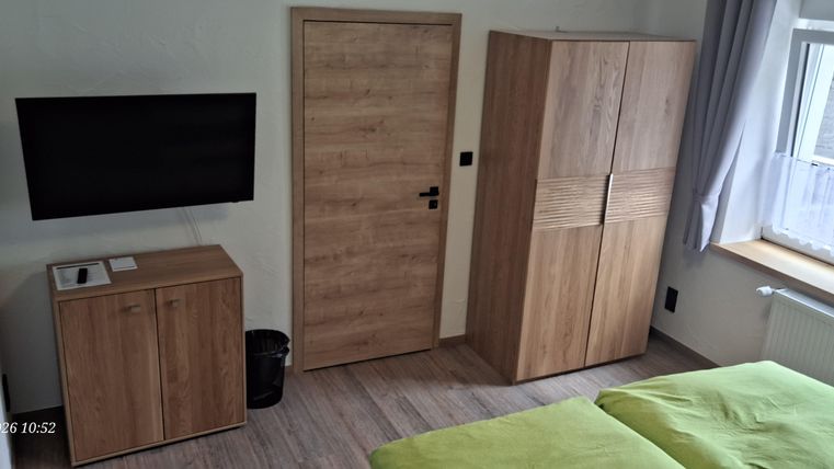 A modern bedroom with a television and a wooden wardrobe. The walls are light and the beds have green bedding.