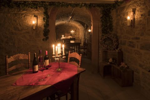 Ein gemütlicher Weinkeller mit Steinmauern und Kerzenlicht. Auf einem Tisch stehen Weinflaschen und eine rote Tischdecke.