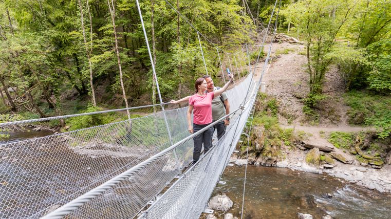 Zwei Personen machen ein Selfie auf einer Hängebrücke über einem Fluss in einem Waldgebiet.