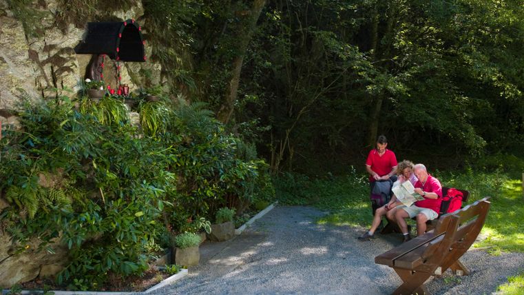 Drei Personen sitzen auf einer Bank im Wald, neben einer Felswand mit einer kleinen Madonna-Statue.