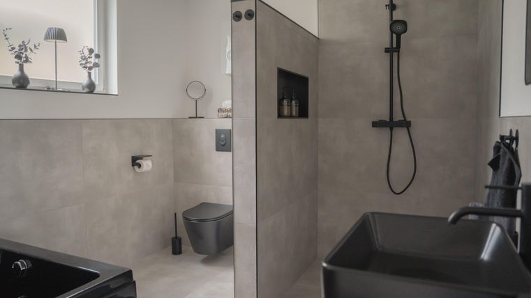 Een moderne badkamer met grijze tegels en elegante zwarte sanitair. De douche is open en goed verlicht, met een raam dat daglicht binnenlaat.