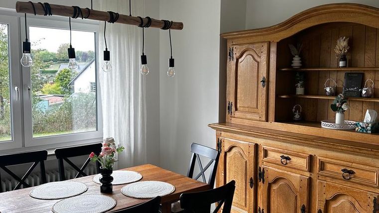 Ein einladender Essbereich mit einem Holz Tisch und vier Stühlen. Eine große Vitrine aus Holz steht neben einem Fenster mit Vorhängen.