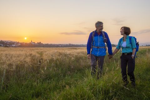 A couple walks through a field at sunset, with a town in the background.