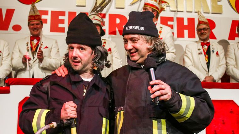 Two men in firefighter uniforms are standing on stage and talking to each other. In the background, other people dressed in festive clothing can be seen.