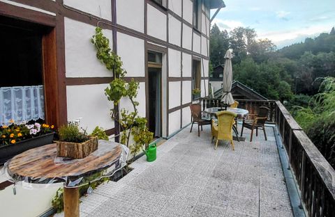 A cozy terrace with a table and chairs, surrounded by plants. In the background, a green landscape can be seen.