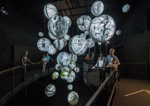 Eine moderne Ausstellung mit schwebenden, beleuchteten Kugeln, die Naturmotive zeigen. Besucher beobachten die Installation in einem dunklen Raum.