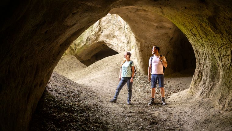 Twee mensen staan in een grote, natuurlijke grot met aardse muren en openingen.