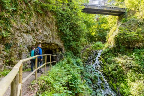 Twee mensen staan voor een grot in een groene, bosrijke omgeving met een kleine waterval en een brug erover.
