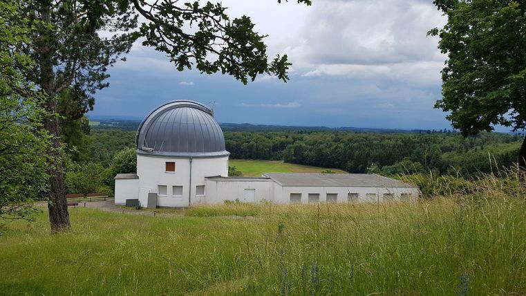 Eine Observatoriumsgebäude inmitten von grünen Wiesen und Bäumen. Der Himmel ist bewölkt, und die Landschaft erstreckt sich über sanfte Hügel.