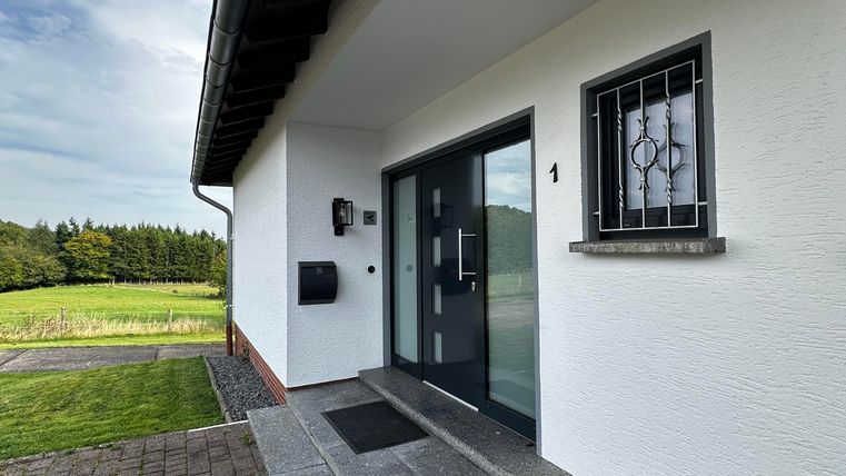 Een moderne entree met een grijze deur en grote ramen. Het pad is ontworpen met klinkers en leidt naar een verzorgd groen gebied.