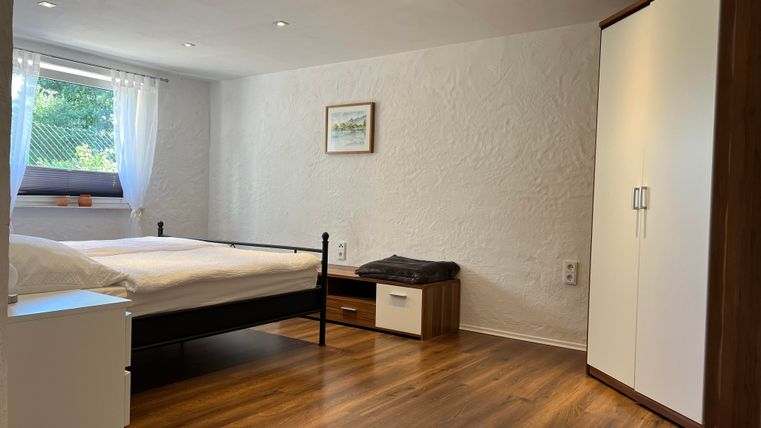 Ein helles Schlafzimmer mit einem Doppelbett und einem großen Fenster. Der Boden ist aus Holz, und es gibt einen Schrank sowie eine kleine Kommode.