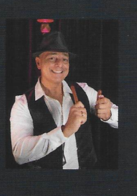 A man in a hat with a smile, who is working on an object with one hand. In the background, a black curtain is visible.