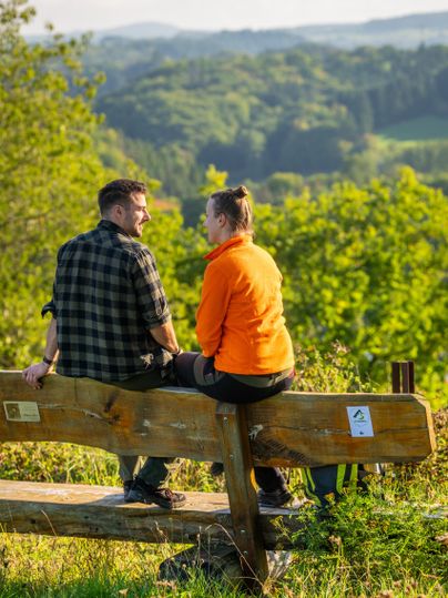 A couple is sitting on a bench and enjoying the view of a green landscape. The sun is shining and the atmosphere feels relaxed.