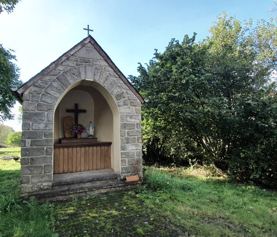 Une petite chapelle en pierre avec une croix et une statue de la Vierge, entourée d'arbres verts et d'herbe., © TI Bitburger Land
