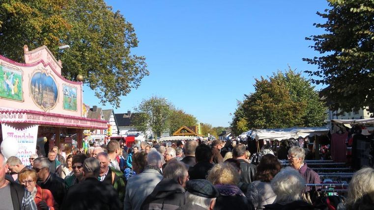 A bustling market with many people. Colorful stalls and autumn trees are visible.