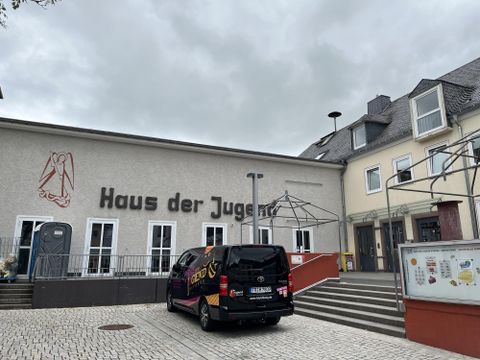Das Bild zeigt das "Haus der Jugend" mit einem großen Schriftzug an der Fassade. Vor dem Gebäude steht ein schwarzes Fahrzeug und es sind einige Treppen im Vordergrund sichtbar.