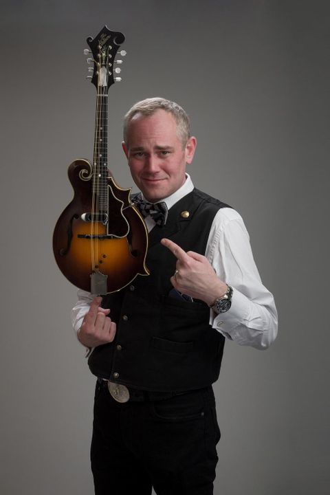 A man is holding a mandolin in his hand and pointing at it with his finger. He is wearing a black vest and a white shirt.