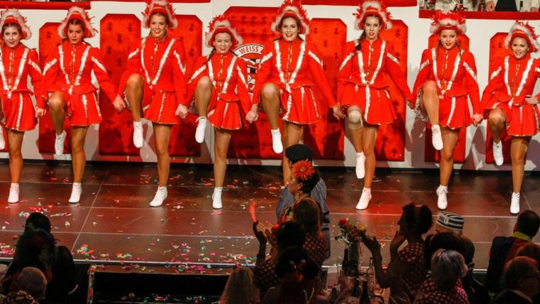 A group of dancers in red costumes is performing a choreography. The stage is festively decorated and the audience is applauding.