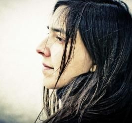 A woman with long, dark hair looks thoughtfully to the side. Her facial expression is friendly and contemplative.