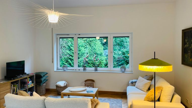 Un salon confortable avec des meubles modernes et de grandes fenêtres. À l'extérieur, on peut voir des plantes vertes.