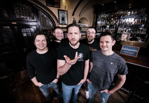 A band is standing in a cozy pub. They are smiling and posing in front of a bar with many drinks.