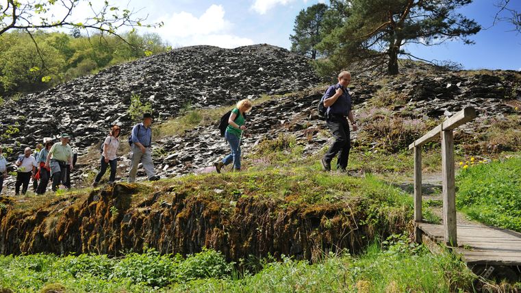 A group of hikers are walking along a narrow path. A steep, dark slate mountain with scattered vegetation can be seen in the background. 