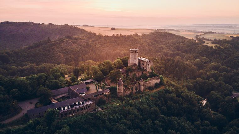 Burg Pyrmont liegt auf einem bewaldeten Hügel, umgeben von sanften Höhenzügen. Die mittelalterliche Anlage mit rundem Bergfried und Ringmauer wird im warmen Licht eines Sonnenuntergangs stimmungsvoll beleuchtet.