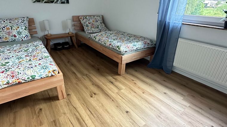 Ein einfaches Schlafzimmer mit zwei Einzelbetten und bunten Bettdecken. Ein großes Fenster mit einem blauen Vorhang sorgt für viel Tageslicht.