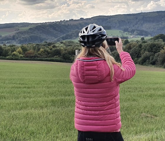 Une personne portant une veste rose et un casque de vélo photographie un paysage vert avec des collines en arrière-plan., © TI Bitburger Land