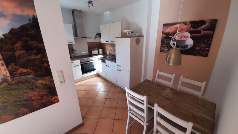 Eine moderne Küche mit Holz-Esstisch und weißen Stühlen. An der Wand sind ansprechende Bilder und die Möbel sind hell und freundlich gestaltet.
