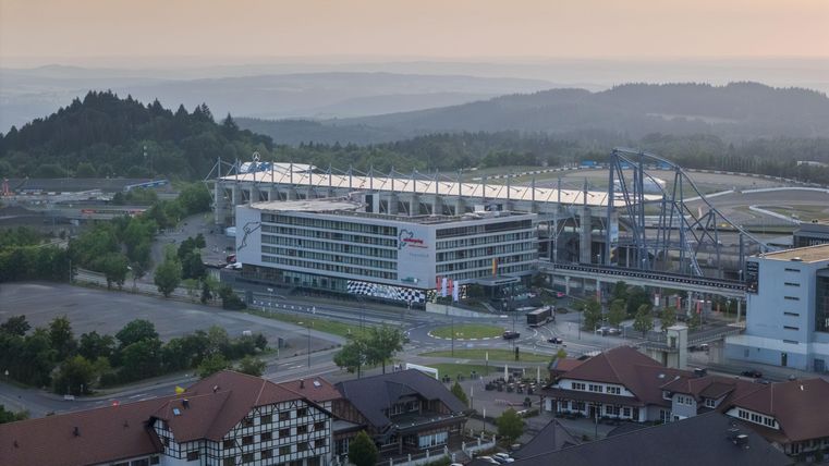 Een uitgestrekt uitzicht op een sportstadion met nabije gebouwen en berglandschap op de achtergrond. De lucht is verlicht in zachte kleuren.