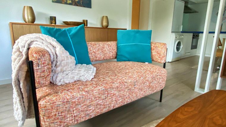 Un canapé confortable avec un tissu coloré et des coussins décoratifs se trouve dans une pièce moderne. En arrière-plan, on peut voir quelques meubles et une salle de bain.