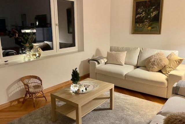 Ein gemütliches Wohnzimmer mit einem hellen Sofa und einem niedrigen Tisch. An der Wand hängt ein Gemälde und vor einem großen Fenster steht ein kleiner dekorativer Baum.