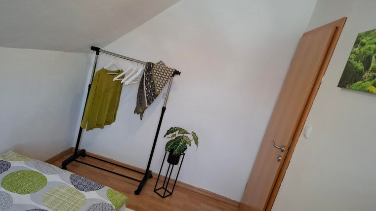 Een lichte kamer met een bed en een kledingstandaard. Aan de muur hangt een groene plantenfoto, en er is een kleine pot met een plant.