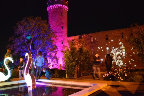 Eine beleuchtete Burg bei Nacht mit bunten Lichtern und fröhlichen Menschen. Im Vordergrund sind dekorative Elemente und ein Wasserbecken zu sehen.