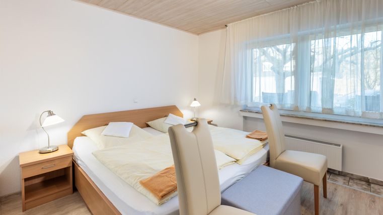 Een eenvoudige slaapkamer met een tweepersoonsbed en lichte meubels. Grote ramen zorgen voor veel natuurlijk licht.