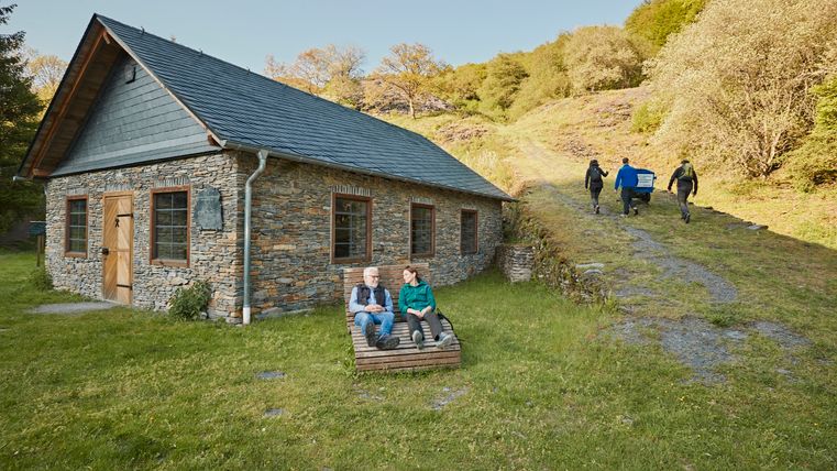 Une maison en pierre pleine de charme, nichée dans un cadre verdoyant. Deux personnes sont assises, détendues, sur un banc, tandis que d'autres s'affairent à l'arrière-plan.
