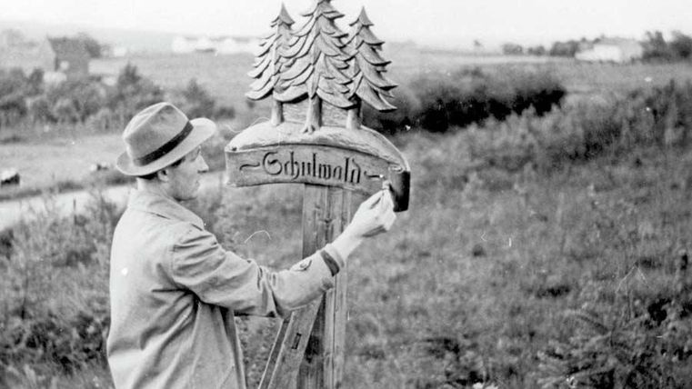 Un homme peint un panneau avec l'inscription "Schlumath". À l'arrière-plan, on peut voir des prairies vertes et des arbres.