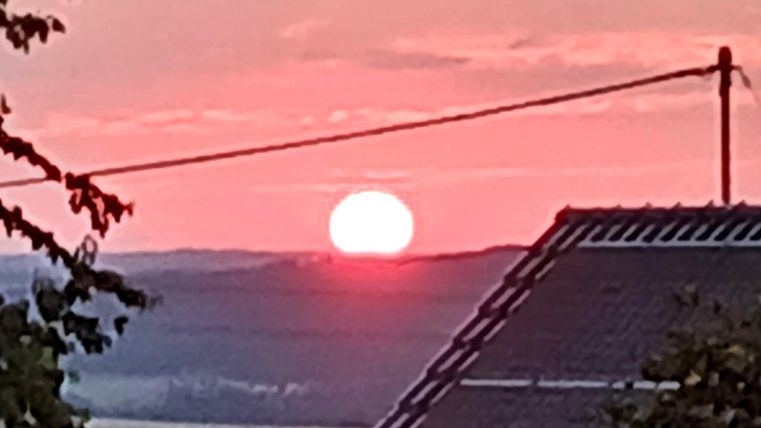 A sunset with a large, glowing sun ball on the horizon. Soft colors of the sky and silhouettes of buildings are visible in the foreground.