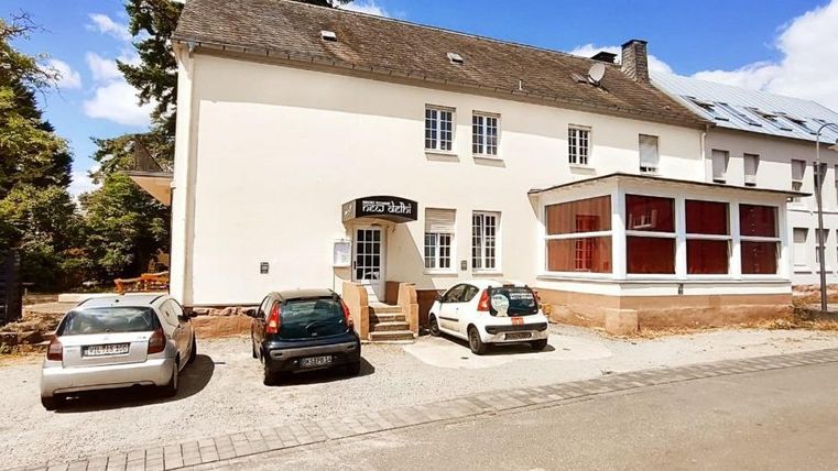 Un bâtiment avec plusieurs fenêtres et une zone couverte. Au premier plan, plusieurs voitures sont sur un parking.