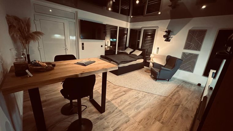 Eine moderne Wohnung mit einem gemütlichen Wohnbereich, einem Esstisch und einem großen Bett. Die Beleuchtung schafft eine warme und einladende Atmosphäre.