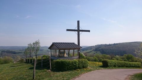 In the middle stands a small covered wooden hut with transparent windows on a hill. It is located at the edge of a blooming meadow of dandelions, which is framed by a belly-high hedge. To the right of the hut stands a tall, brown wooden cross that towers about 3 meters over the hut. On a cloudless sky, one can see far into the Kyll Valley.