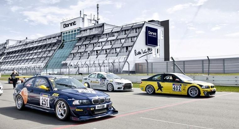 Un groupe de voitures de course circule sur une piste de course. En arrière-plan, le bâtiment du centre de course est visible.