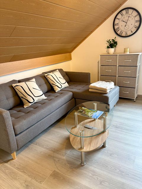 A modern living room with a gray couch and cushions. A round glass table stands in the middle, and a wall decoration with a clock is visible.