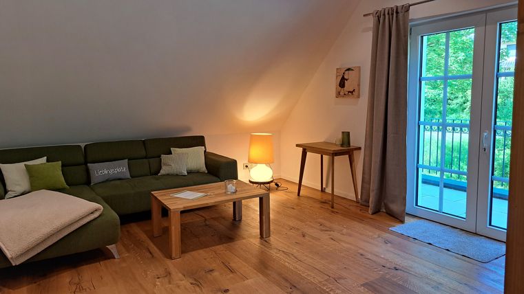 Eine gemütliche Wohnung mit einem grünen Sofa und einem Holztisch. Große Fenster bieten einen Blick auf die grüne Umgebung.