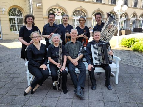 Eine Gruppe von Musikern sitzt auf einer Bank im Freien. Sie spielen verschiedene Instrumente und zeigen Freude an ihrem Zusammenspiel.