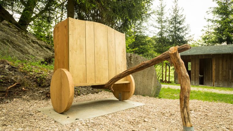 Houten sculptuur van een wagen op een bospad met bomen op de achtergrond.