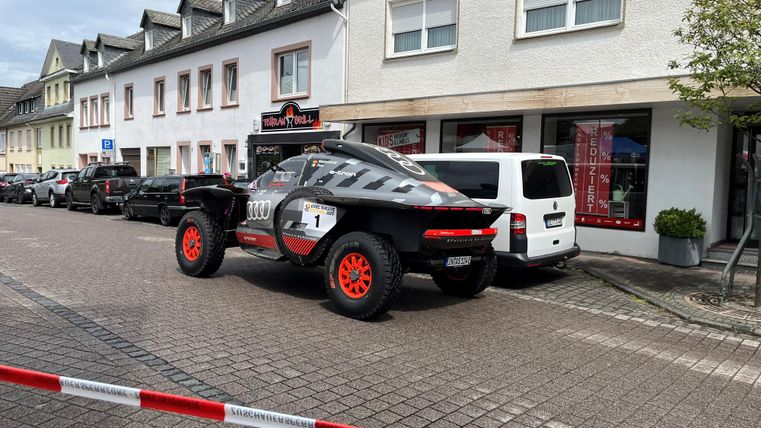 Ein auffälliges Rennfahrzeug steht auf einer Straße vor mehreren Häusern. Im Hintergrund sind weitere Autos und ein Ladengeschäft zu sehen.