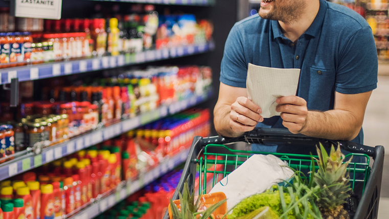 <p>Un homme avec un chariot de courses bien rempli marche dans un rayon du supermarché. Sur les étagères, on peut voir des sauces et du ketchup. Dans sa main, il tient une liste de courses.</p>